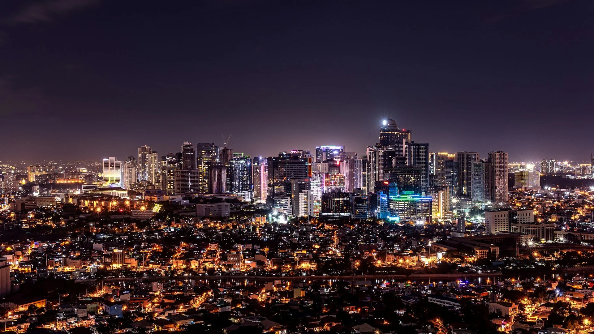 A sprawling cityscape at night, featuring a dense cluster of illuminated skyscrapers against a dark sky.