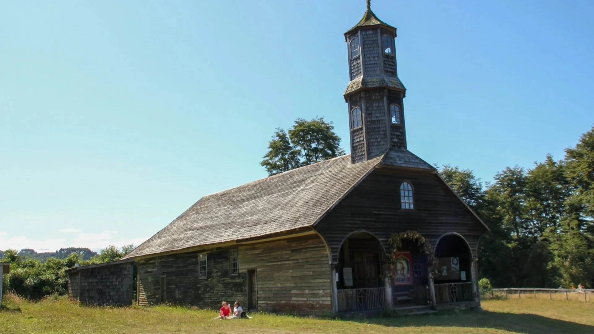 A rustic, multi-tiered wooden church with a shingled roof under a clear blue sky on Chiloé Island.