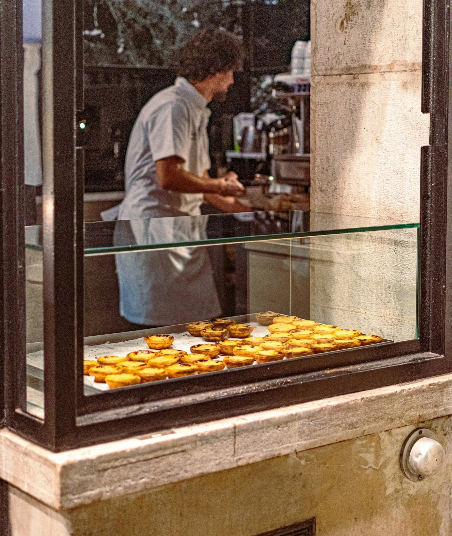 A bakery display window featuring rows of freshly baked Pastéis de Nata, with a baker visible in the background.