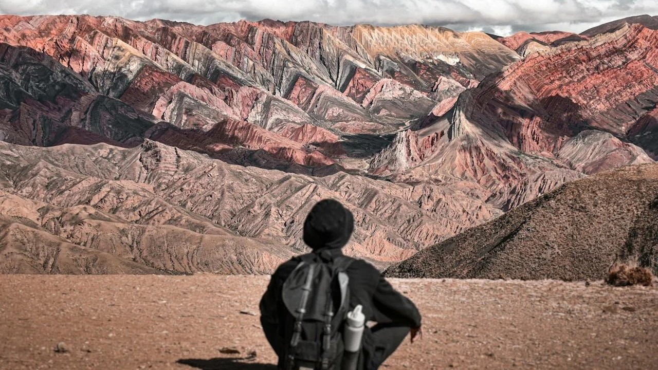 A hiker wearing a backpack sits on a dirt hill from behind, looking out at the stunning, multi-colored ridges of a vast, layered mountain range under a cloudy sky.