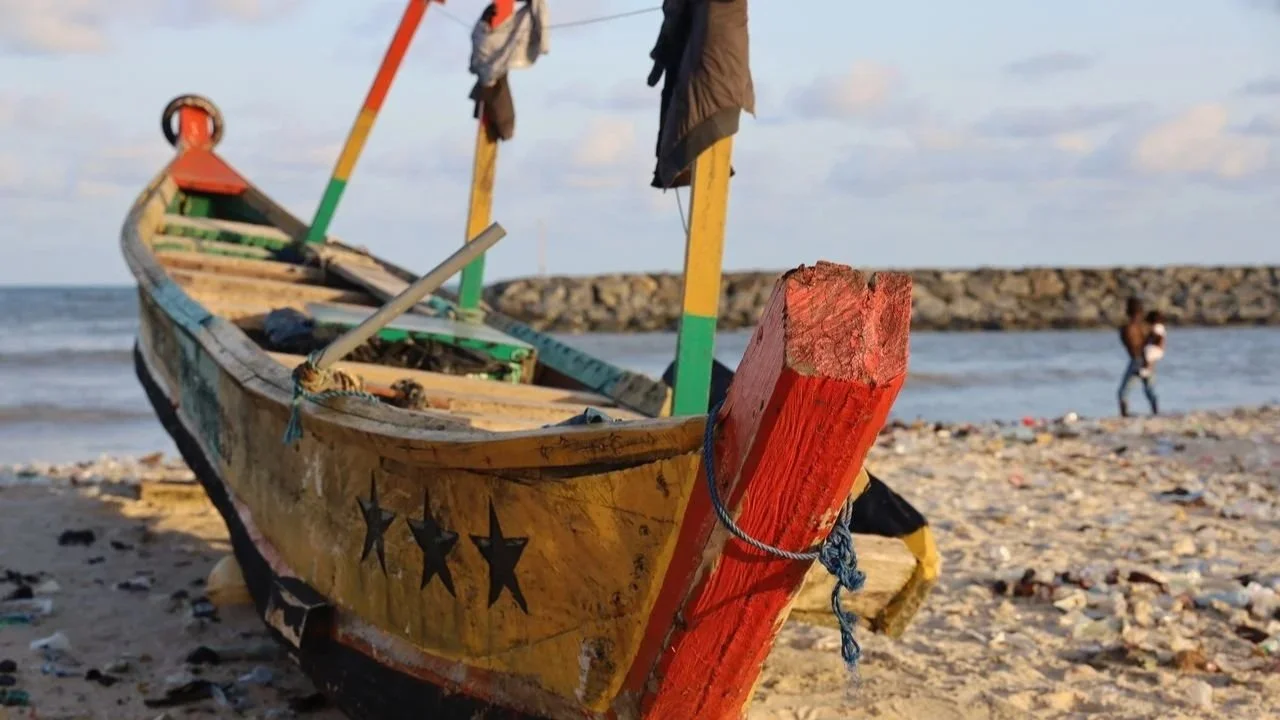 A close-up view of a traditional, brightly painted wooden pirogue boat resting on a sandy beach under a cloudy sky.