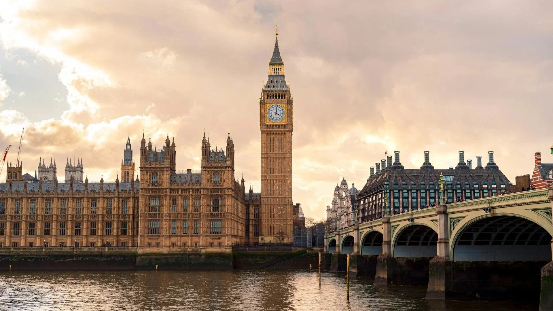 A wide shot of the Palace of Westminster and Big Ben against a golden sunset sky, with Westminster Bridge and the River Thames in the foreground.
