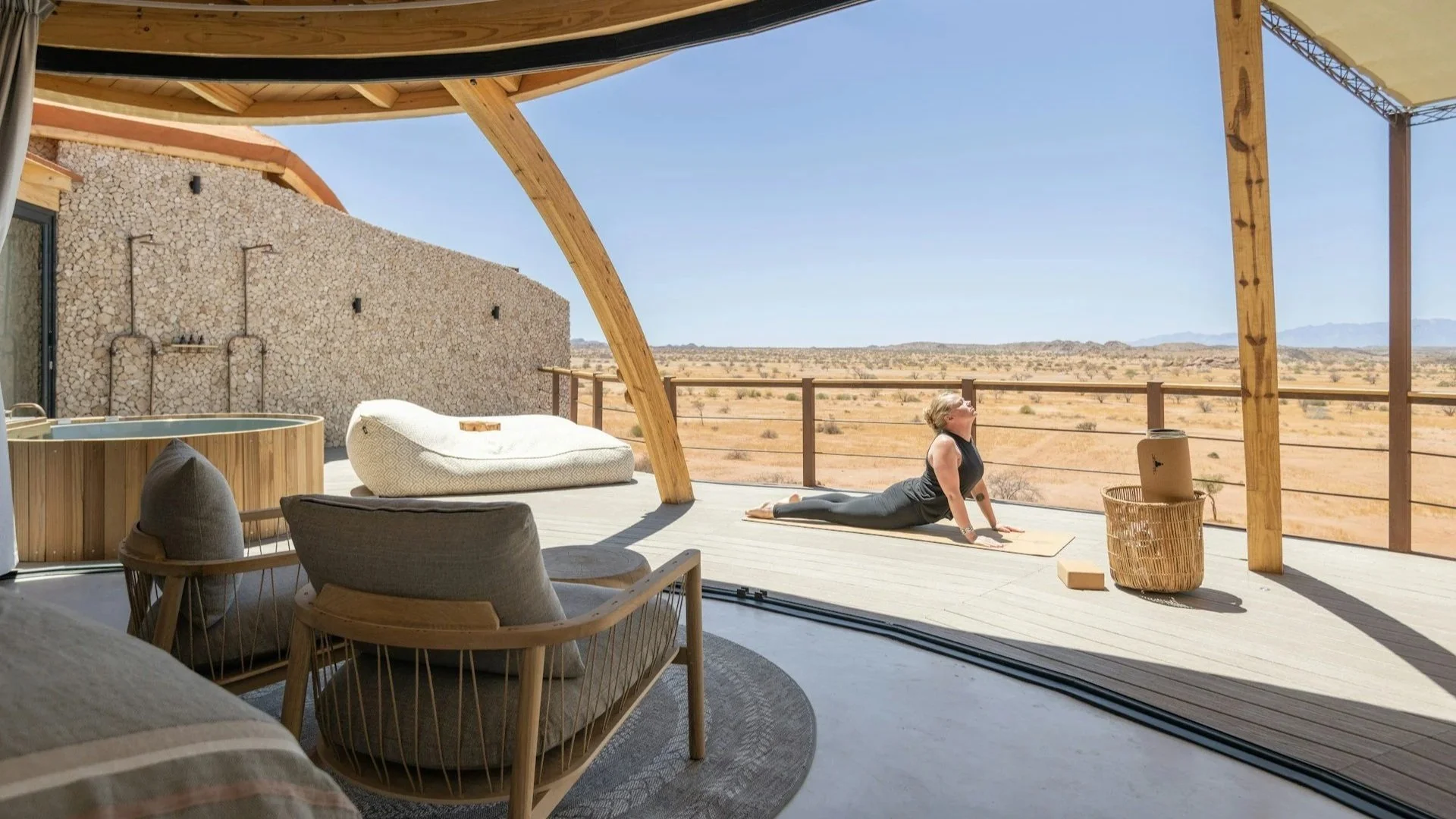 Gemini said A person practices yoga in an upward-facing dog pose on a spacious wooden deck. The deck belongs to a luxury desert lodge with a curved wooden roof.