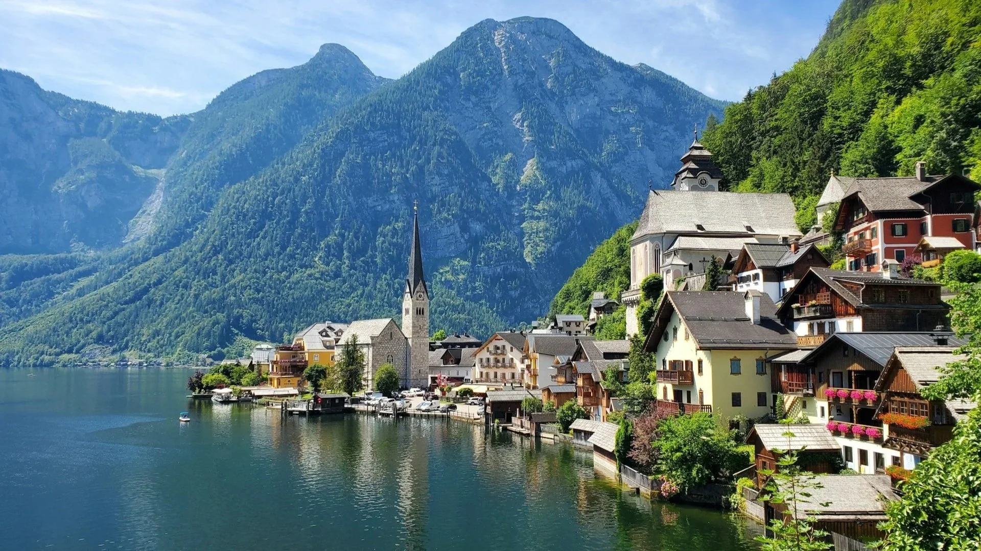 The scenic village of Hallstatt, Austria, featuring colorful houses and a church spire along a calm lake with steep green mountains in the background.