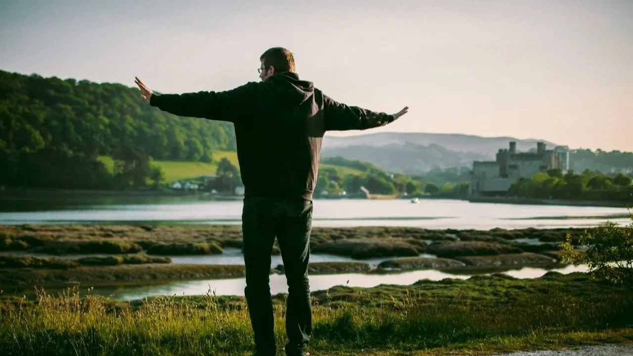 A man standing with his arms outstretched, facing a scenic river landscape with green hills and a castle in the distance.