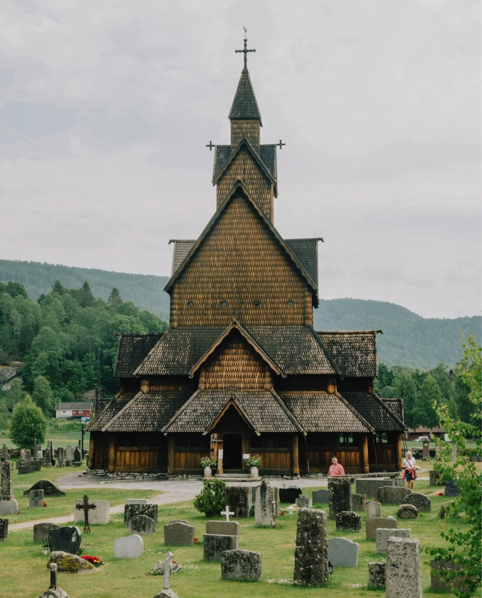 Front view of the historic Heddal Stave Church in Norway, a tiered wooden structure with intricate shingled roofs and crosses, set behind a grassy cemetery under a cloudy sky.