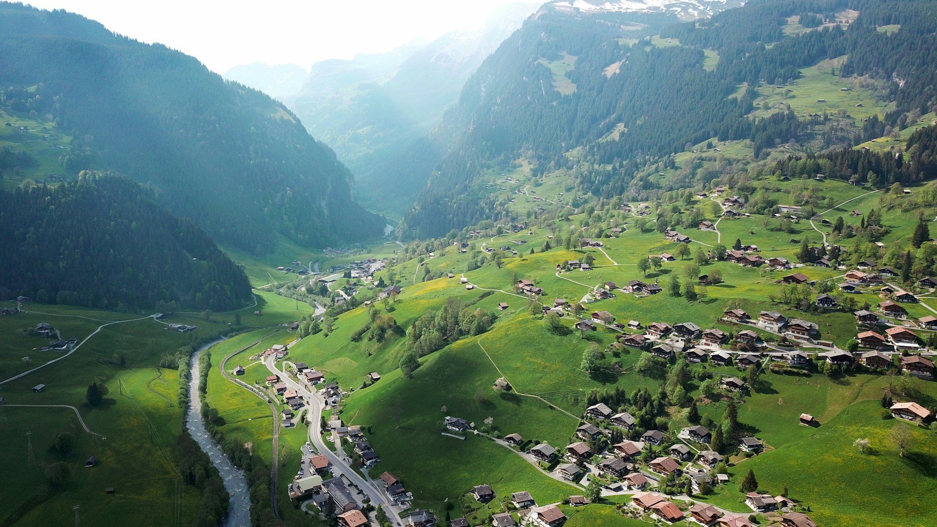An aerial view of a verdant Swiss valley dotted with wooden houses and a winding river, flanked by steep, sunlit mountains.