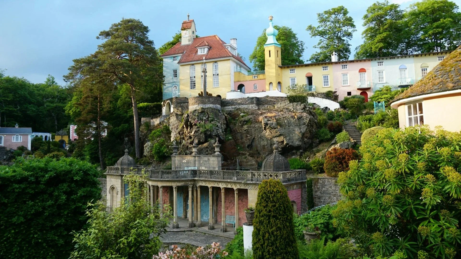 A picturesque village hillside featuring colorful, European-style buildings nestled on rocky cliffs and surrounded by lush green trees.