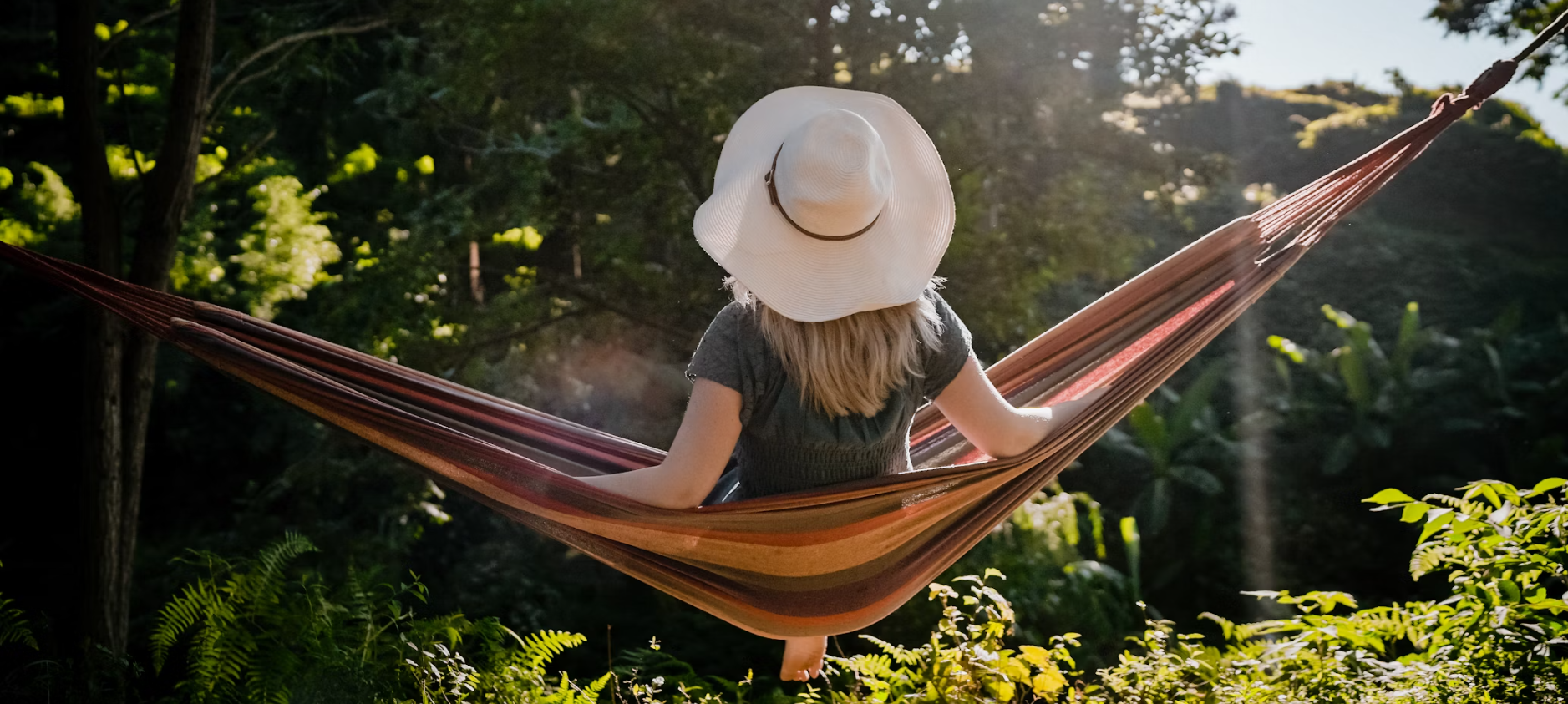 Woman wearing a white sun hat relaxing in a striped hammock in a sunny, green forest.