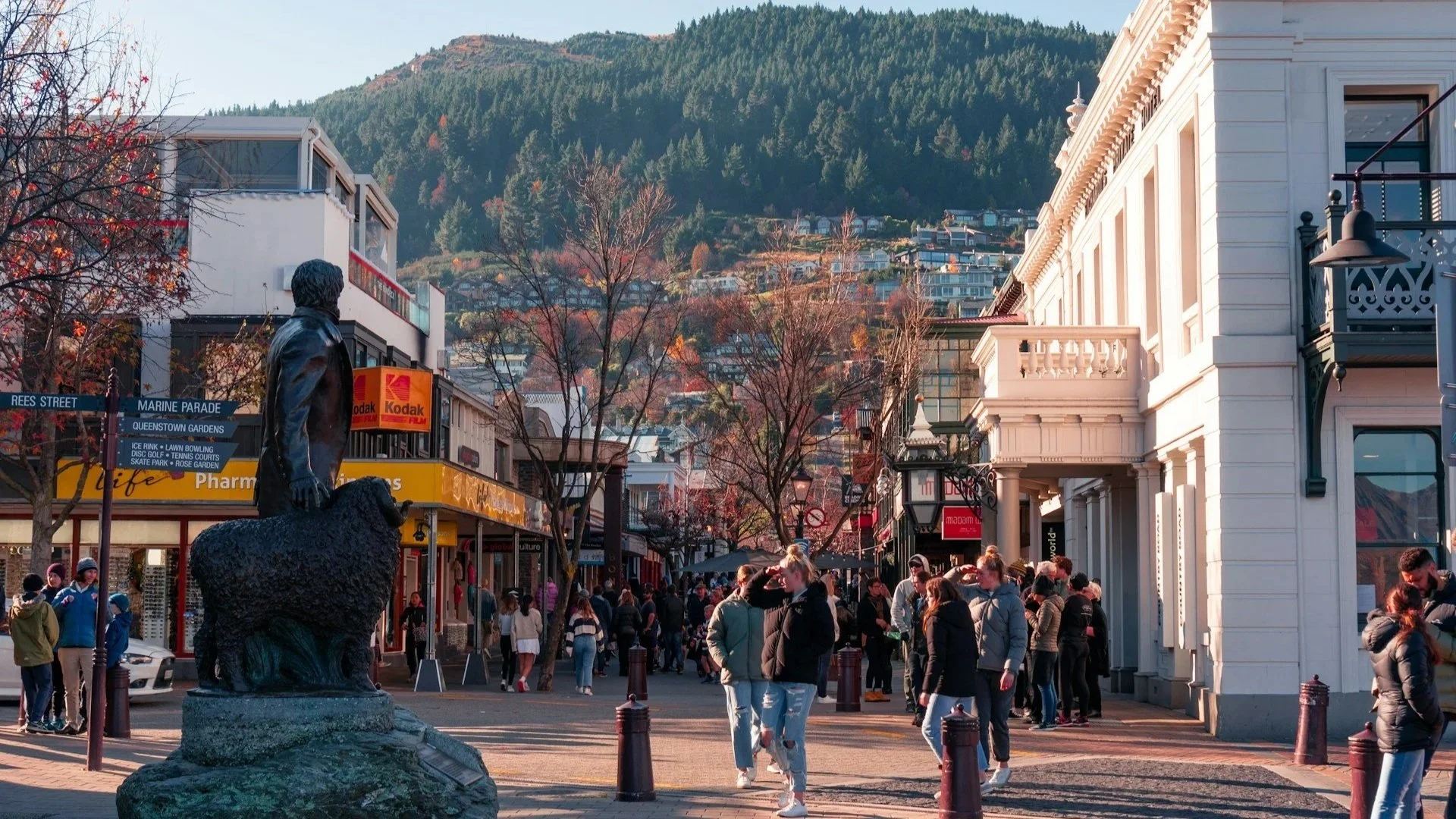 A busy pedestrian street in Queenstown featuring a statue of a man and a sheep against a hillside backdrop.