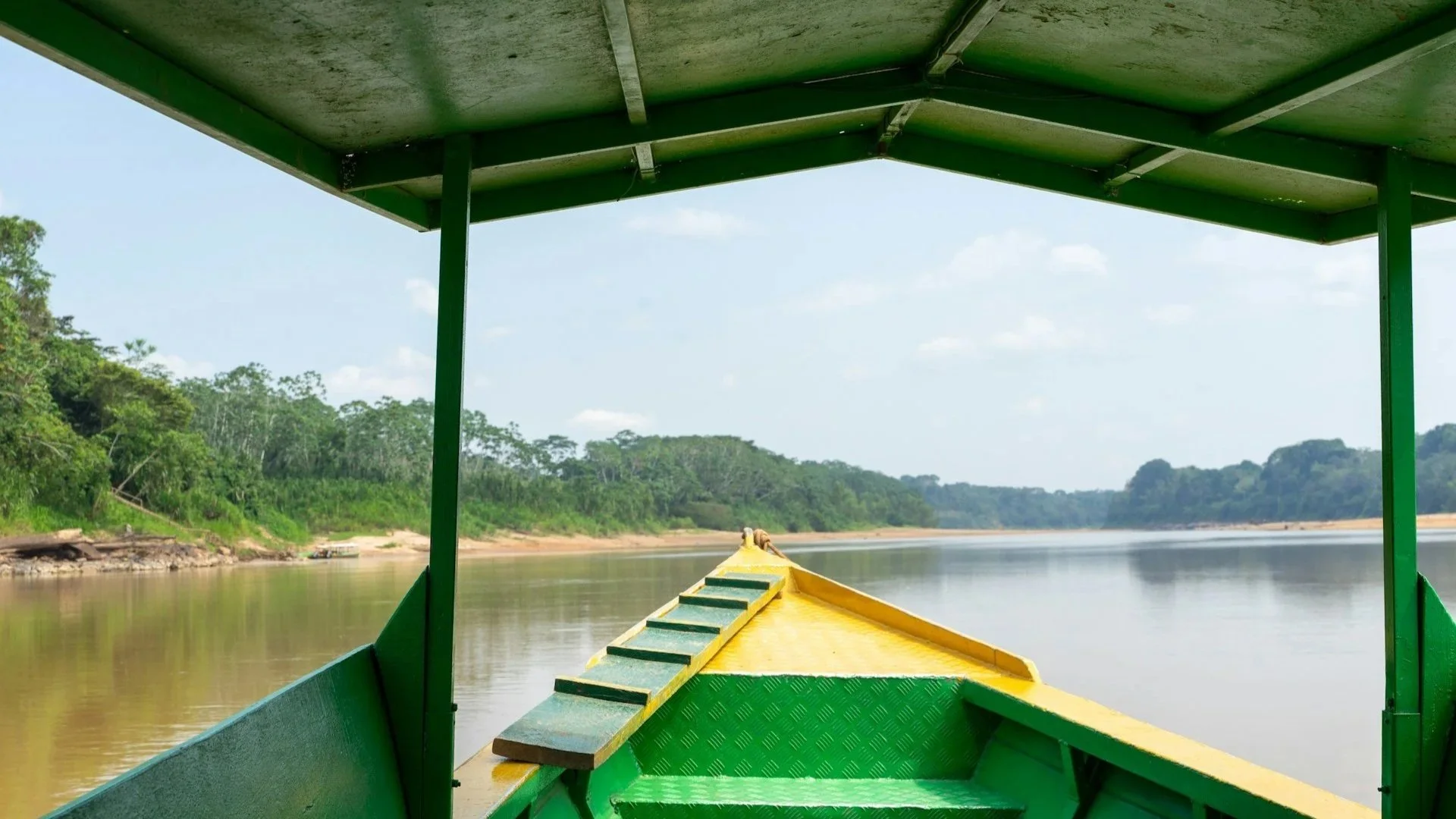 View from the front of a green and yellow motorized boat traveling down a calm river in the Amazon rainforest.