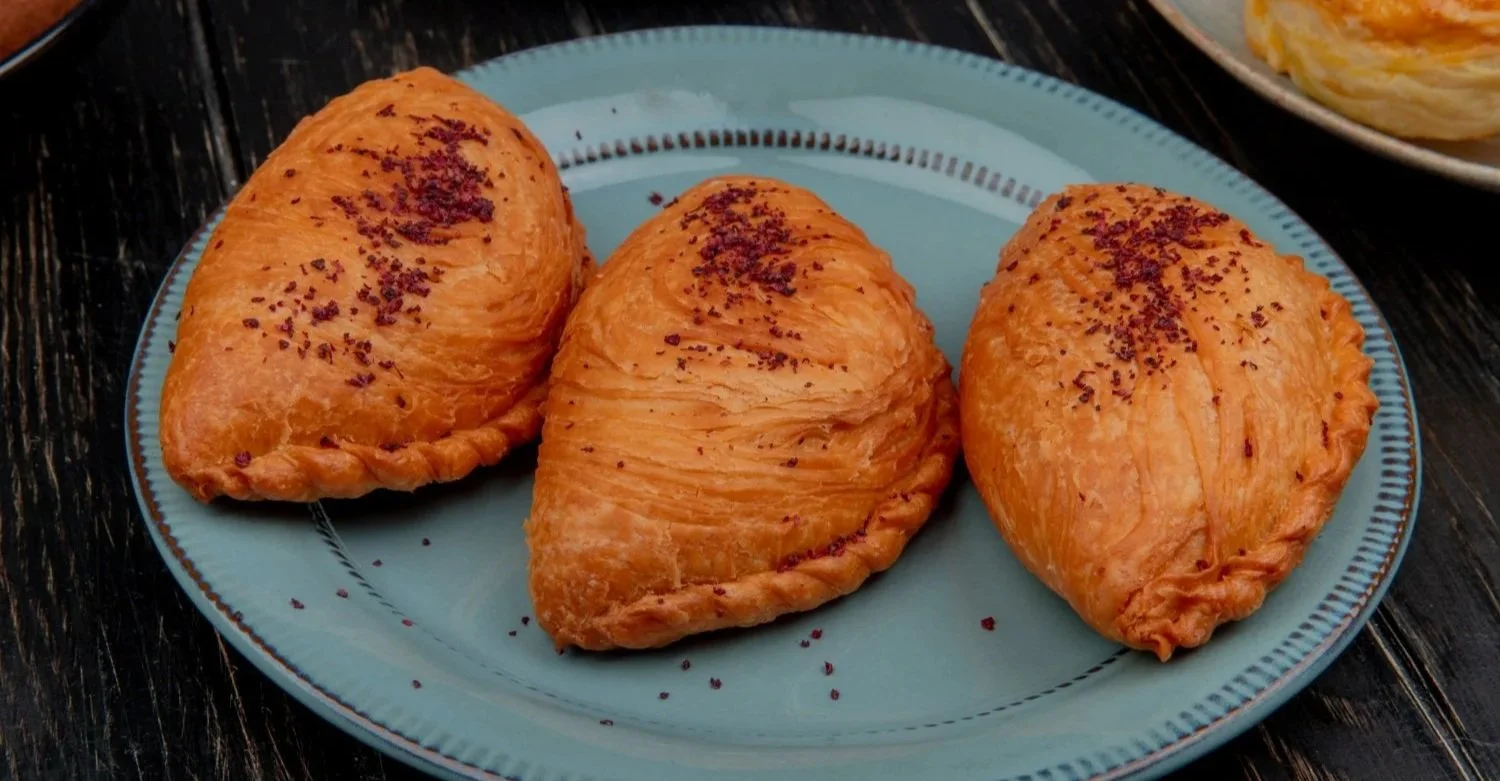 Three golden-brown, flaky pastries dusted with purple spice, arranged on a light blue ceramic plate.