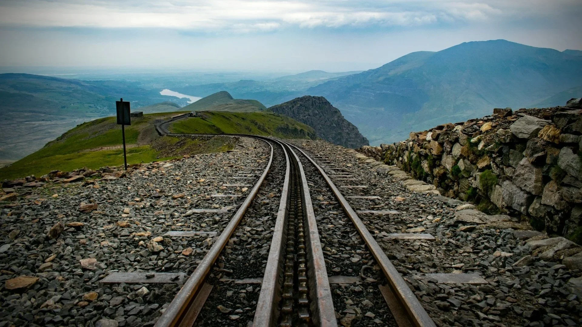 A rack railway track curves along a rocky mountain ridge under a cloudy sky, looking down onto green hills and a distant lake in a valley.