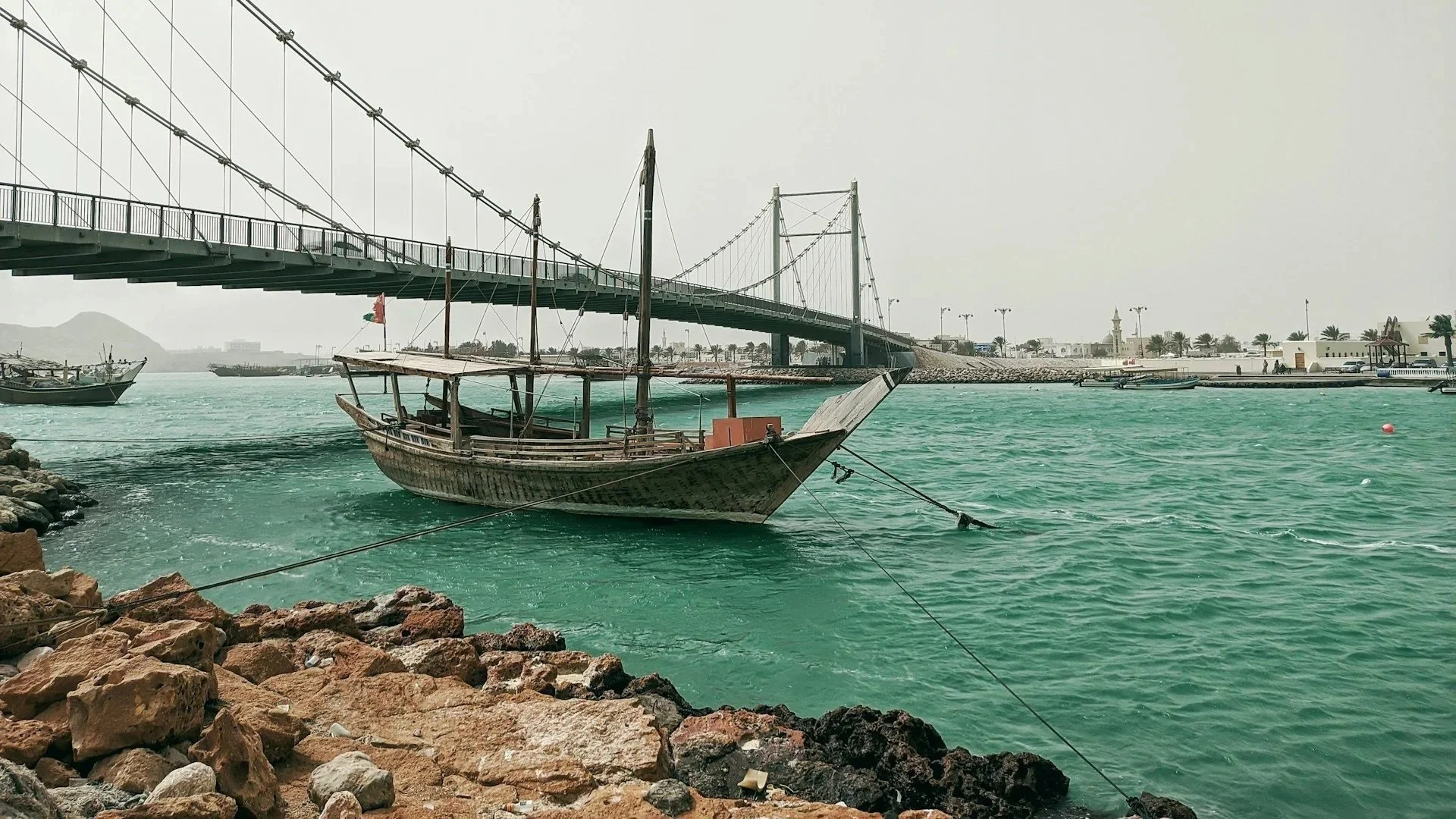 A traditional wooden dhow boat moored in turquoise water beneath a large suspension bridge, with a rocky shoreline in the foreground.