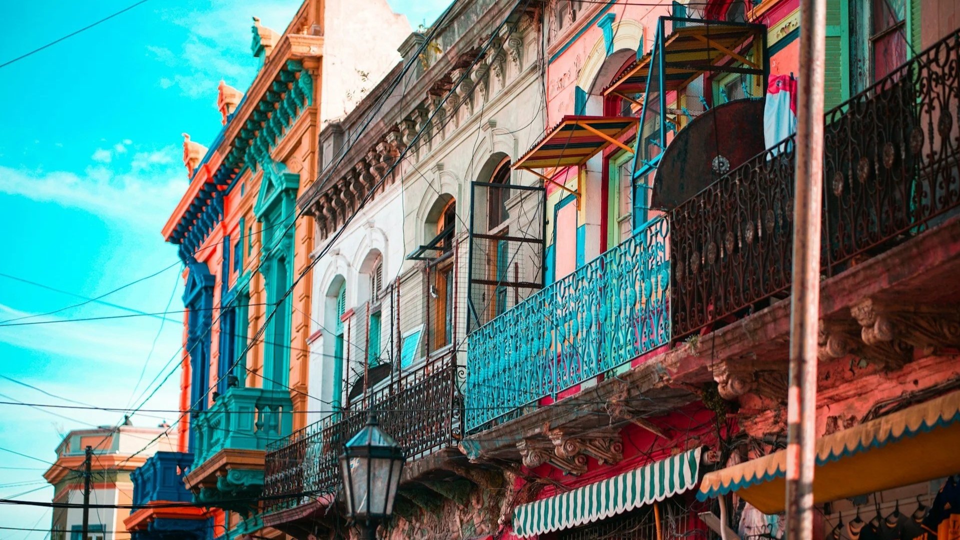 A low-angle view of vibrant, multi-colored buildings with ornate balconies, awnings, and overhead wires under a bright blue sky.