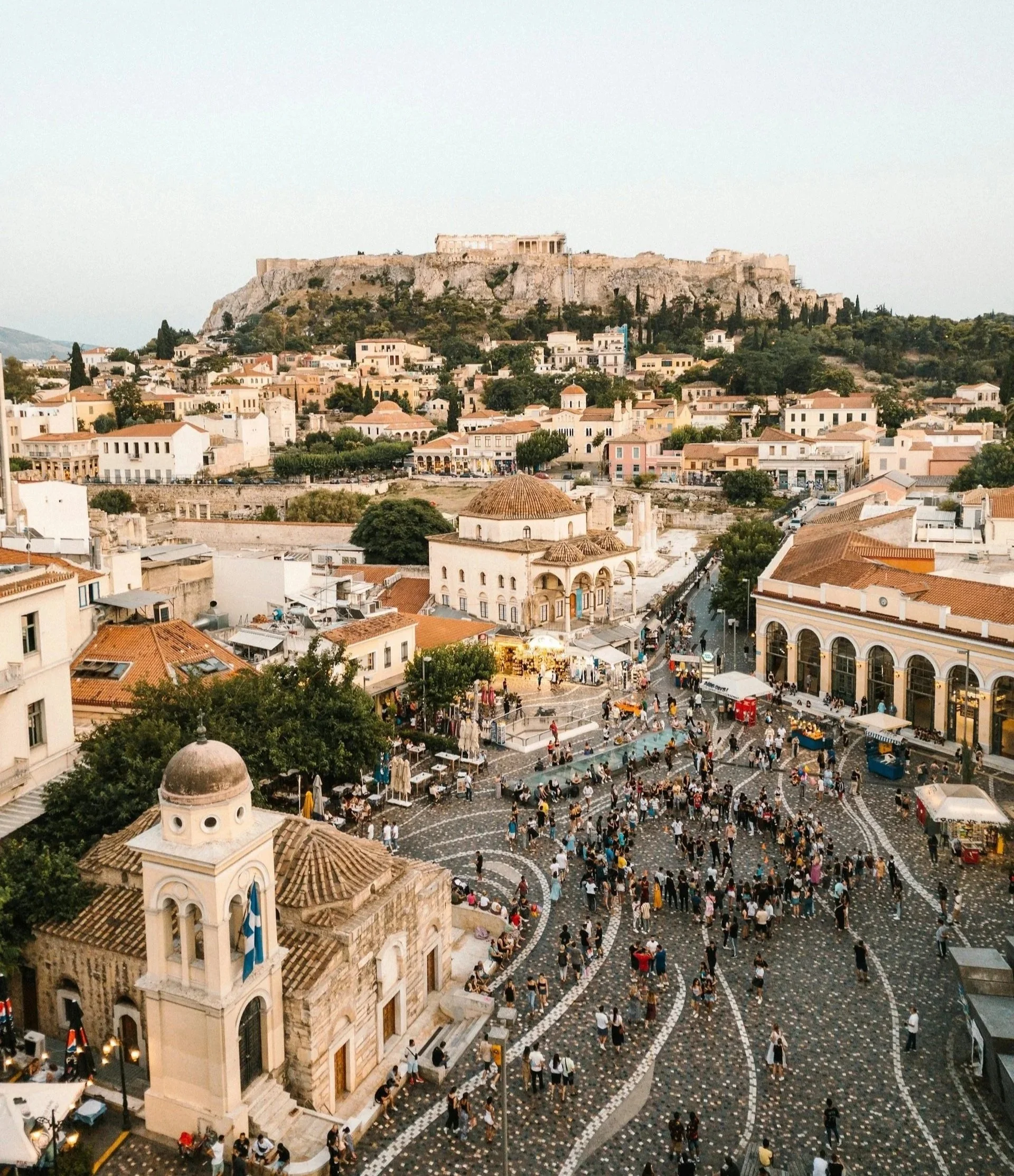 Elevated view of the Plaka district in Athens, Greece, showing the densely packed city buildings and the Acropolis towering on the hill in the background.