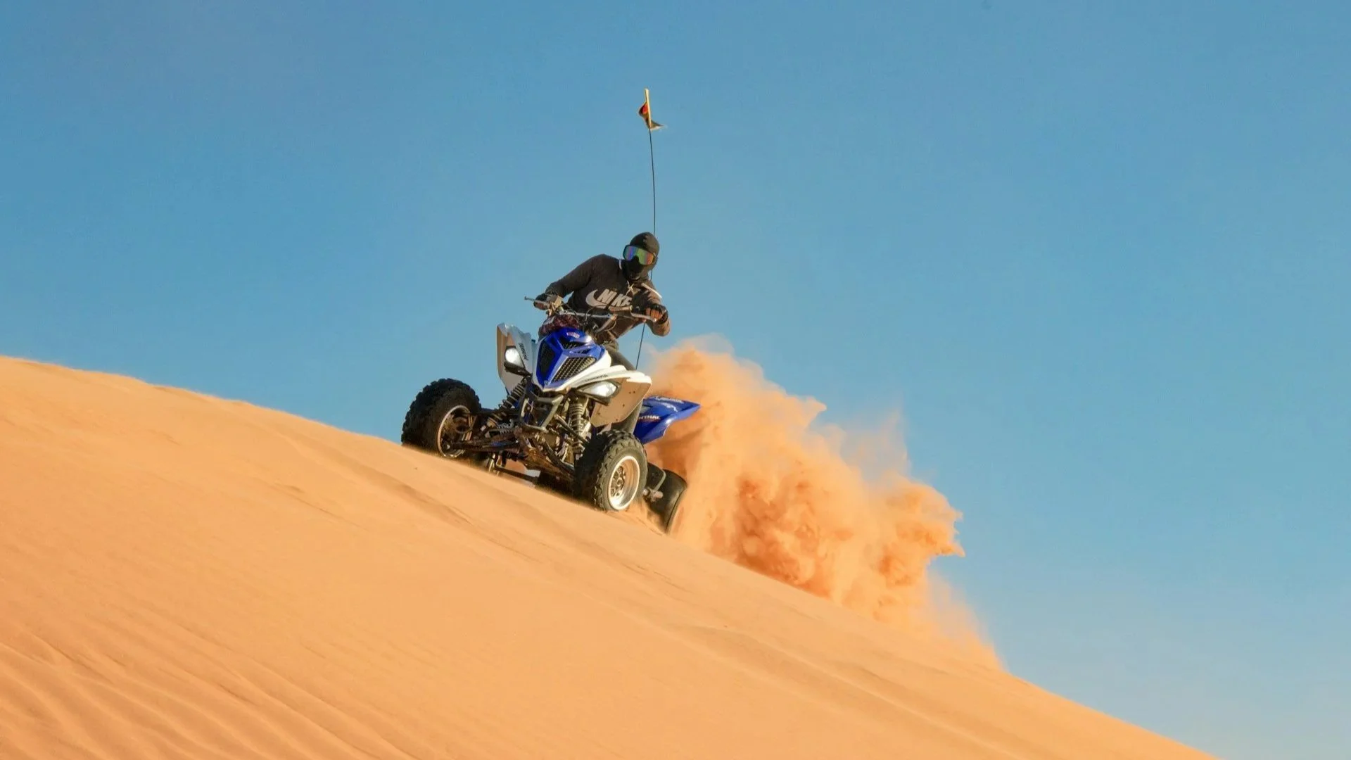 An action shot of a person riding a blue and white quad bike up a steep, orange sand dune under a bright blue sky, kicking up a large cloud of desert dust.