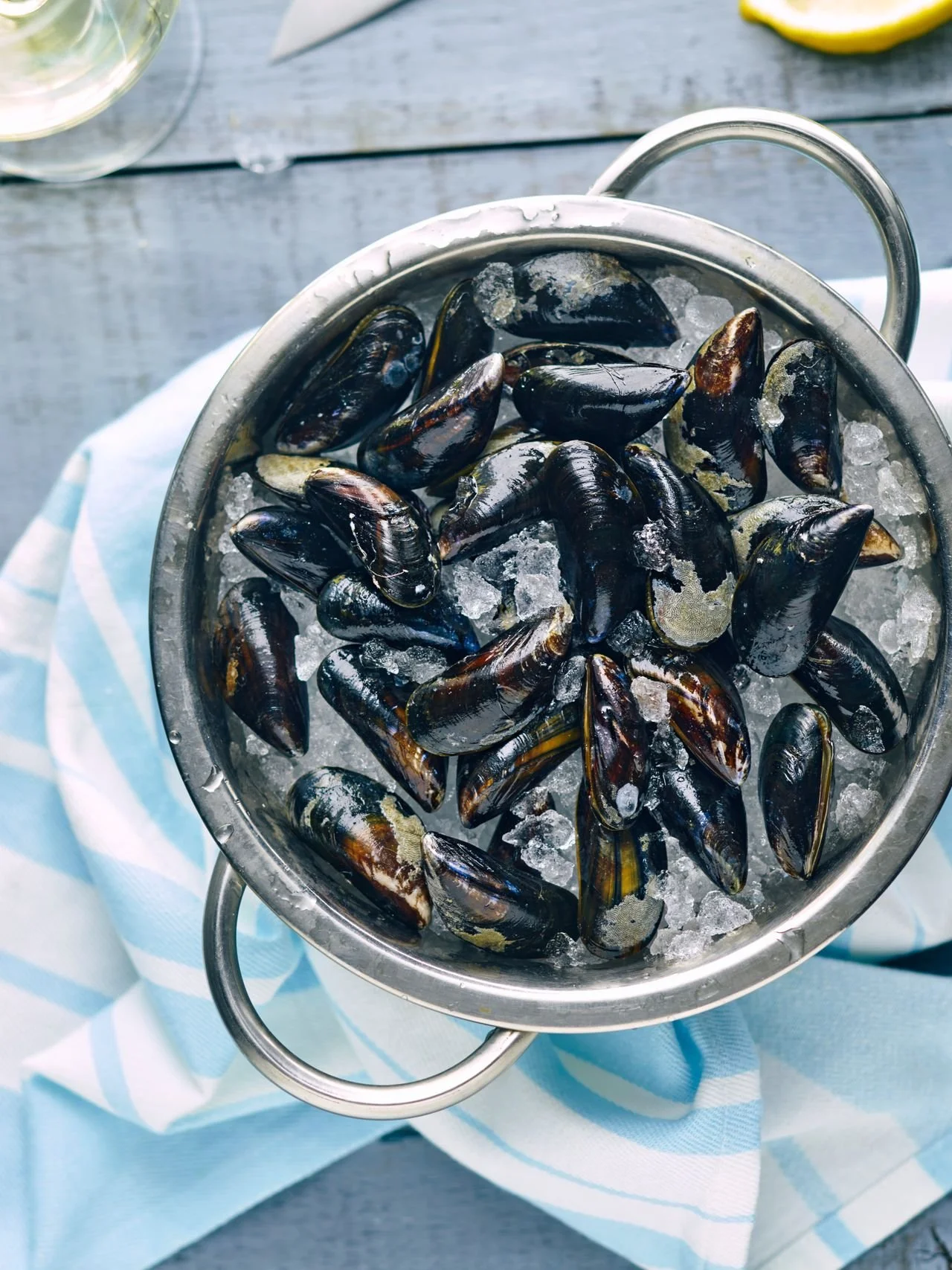 A top-down view of fresh, dark mussels resting on a bed of crushed ice inside a stainless steel colander, placed on a blue and white striped cloth.