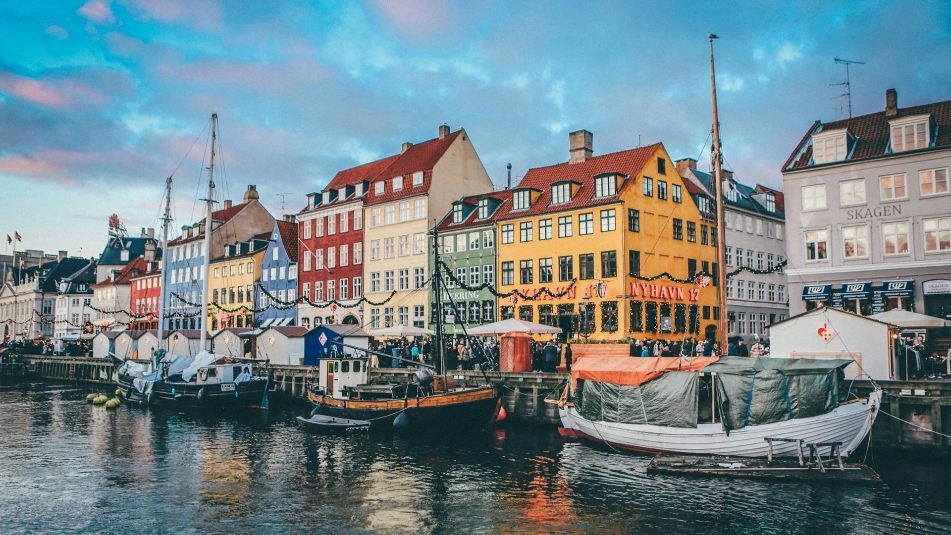 Colorful historic buildings along a canal in Nyhavn, with old wooden boats docked in the water under a twilight sky.