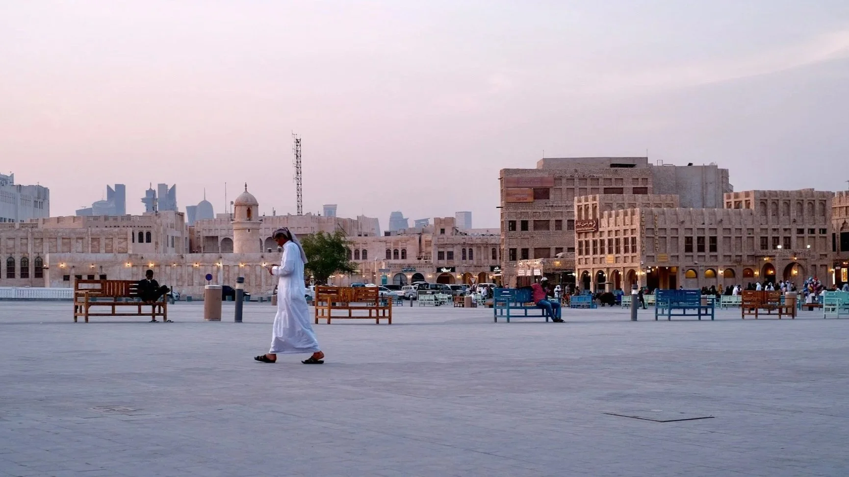 A man wearing a traditional white thobe walks across a wide, open plaza in Souq Waqif at dusk, with the Doha skyline in the far background.
