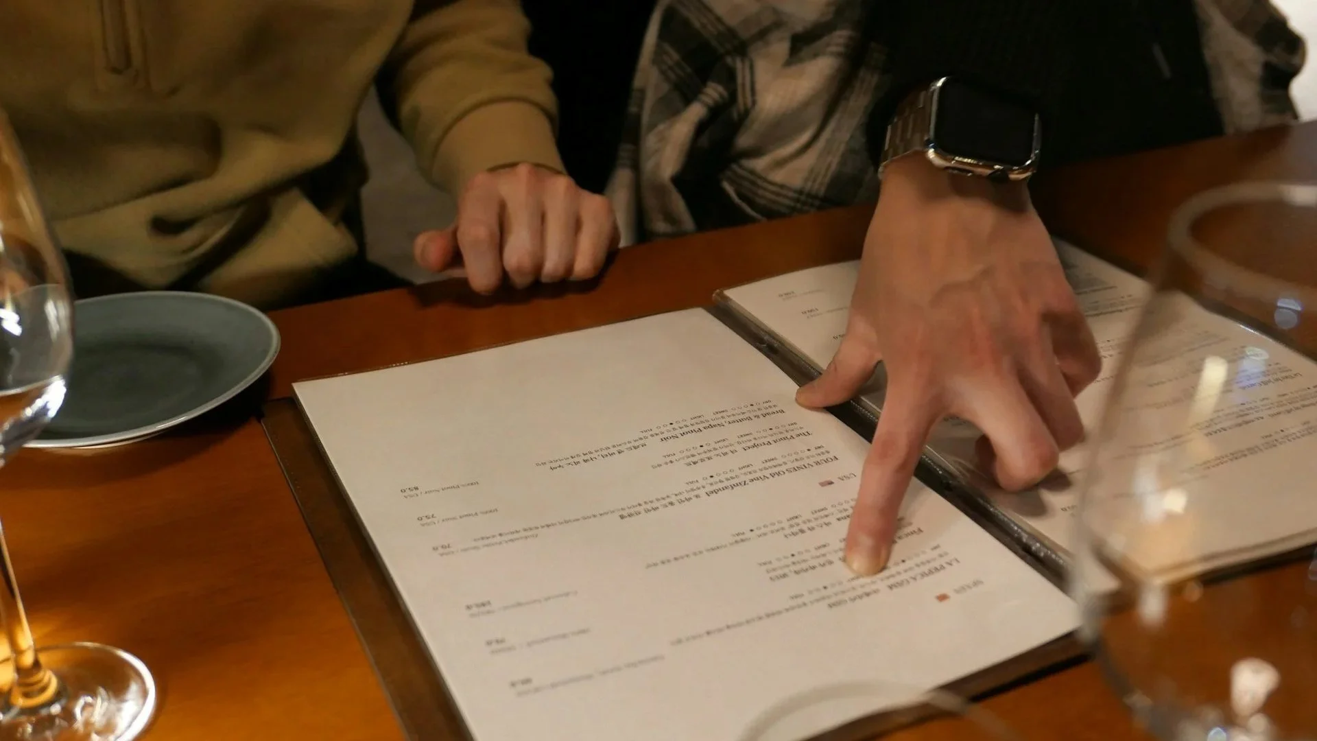 A person’s hand pointing to a specific item on a restaurant menu while sitting at a wooden table with wine glasses and another person nearby.