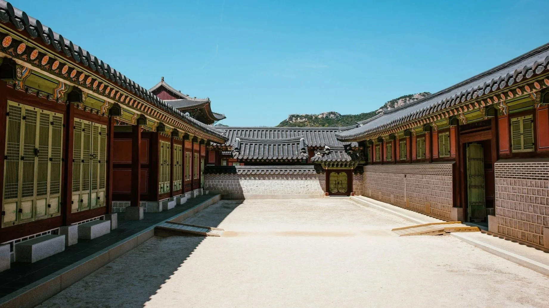 A wide view of a quiet, sunlit courtyard at a Korean palace, surrounded by historic wooden buildings with intricate tiled roofs.