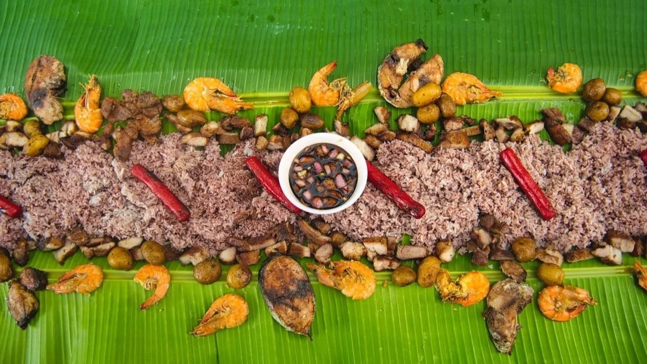 A traditional Filipino boodle fight feast spread across large green banana leaves, featuring a long row of rice topped with grilled fish, shrimp, pieces of pork, red hot dogs, and a central bowl of dipping sauce.