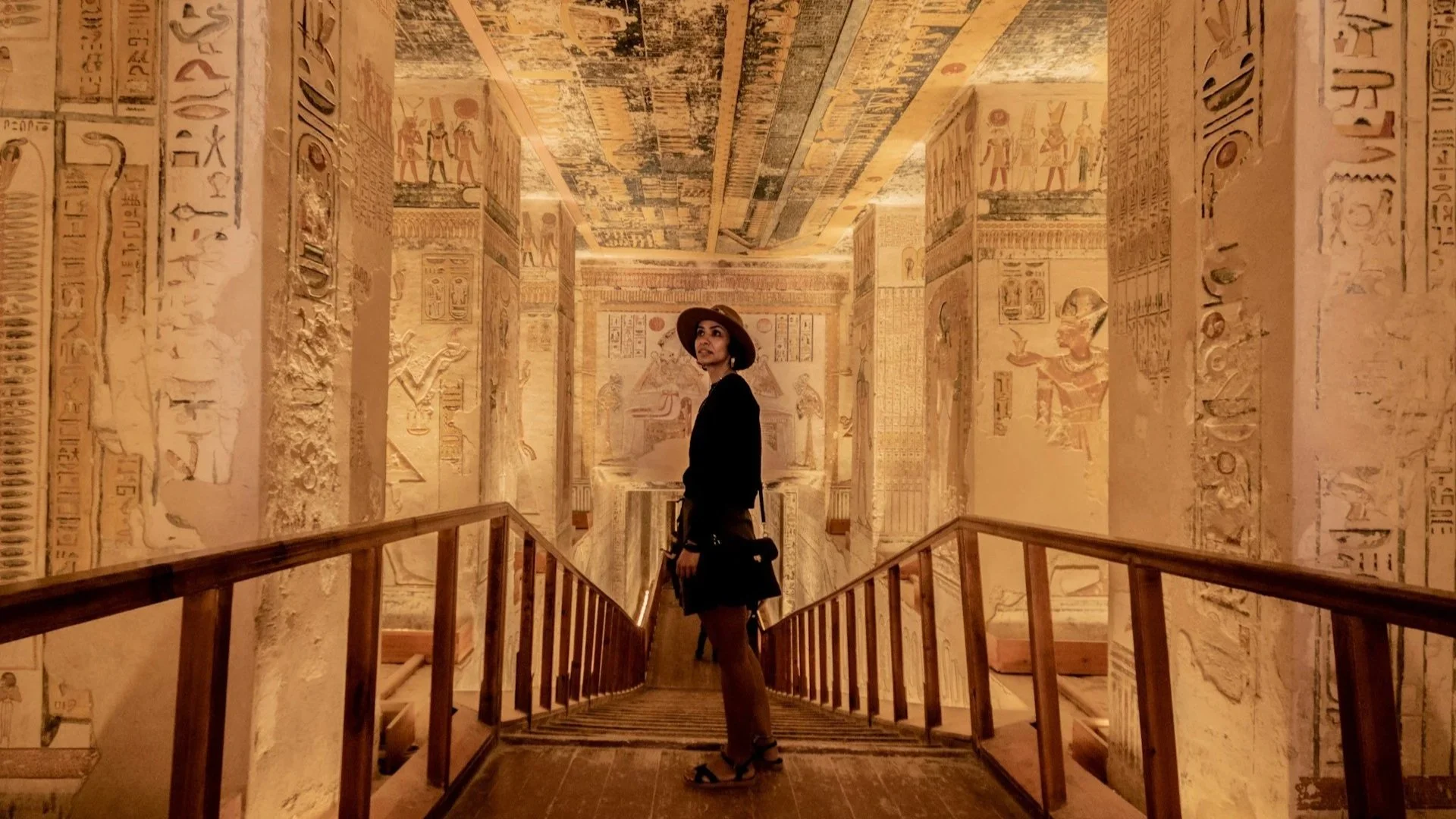 Gemini said A person wearing a wide-brimmed hat and black clothing stands on a wooden staircase leading down into an ancient Egyptian tomb.