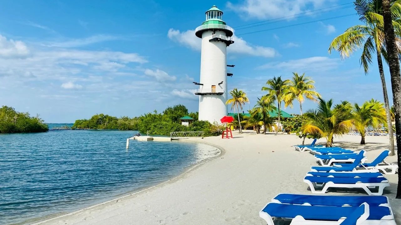 A tall white lighthouse with a green top stands on a sandy beach lined with palm trees and blue lounge chairs under a bright blue sky.