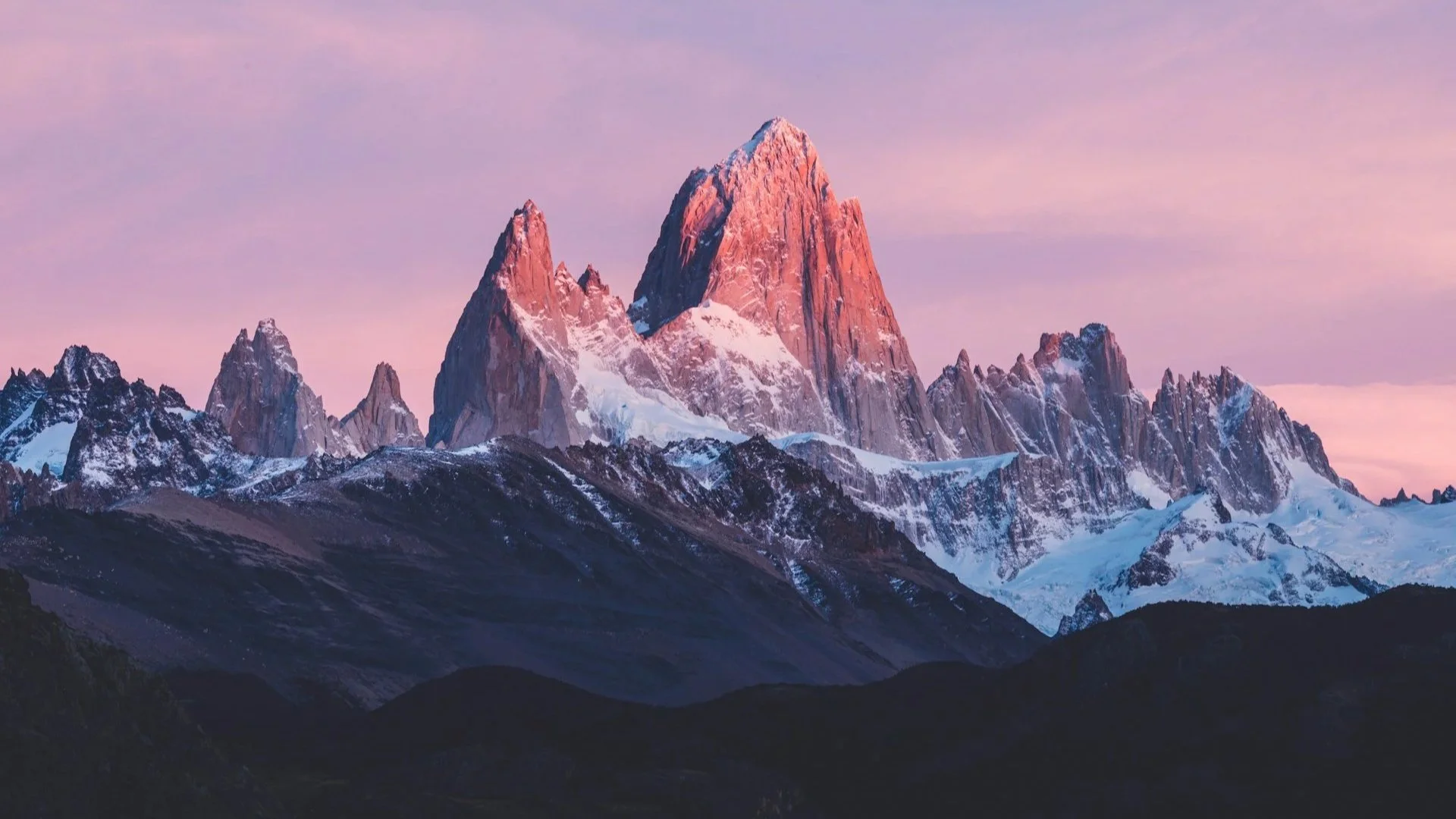 Jagged mountain peaks partially covered in snow, illuminated by a pink and purple glow from a sunset or sunrise sky.