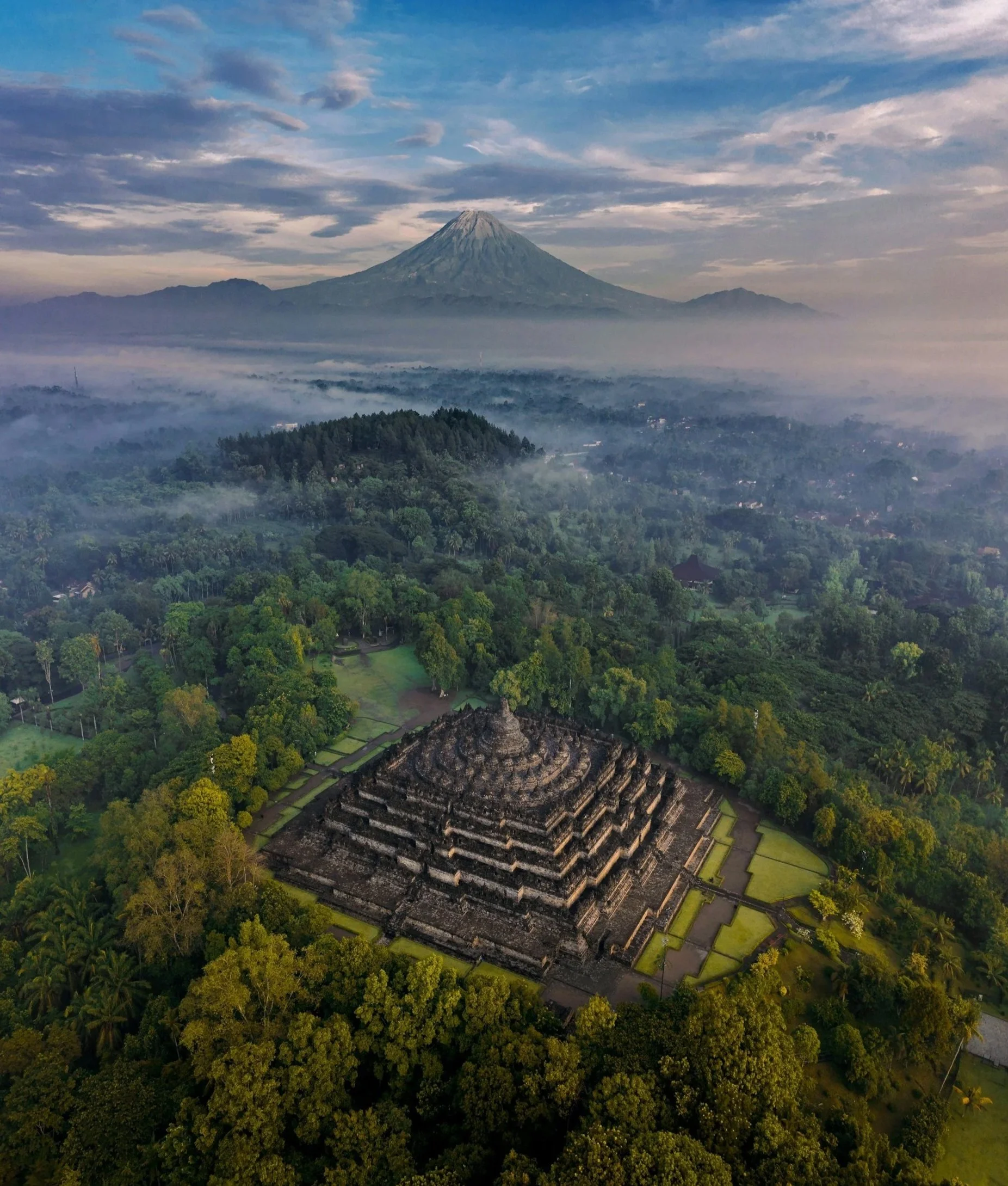 A high-angle view of the Borobudur Buddhist temple nestled in a lush green forest with a mountain peak in the distance.