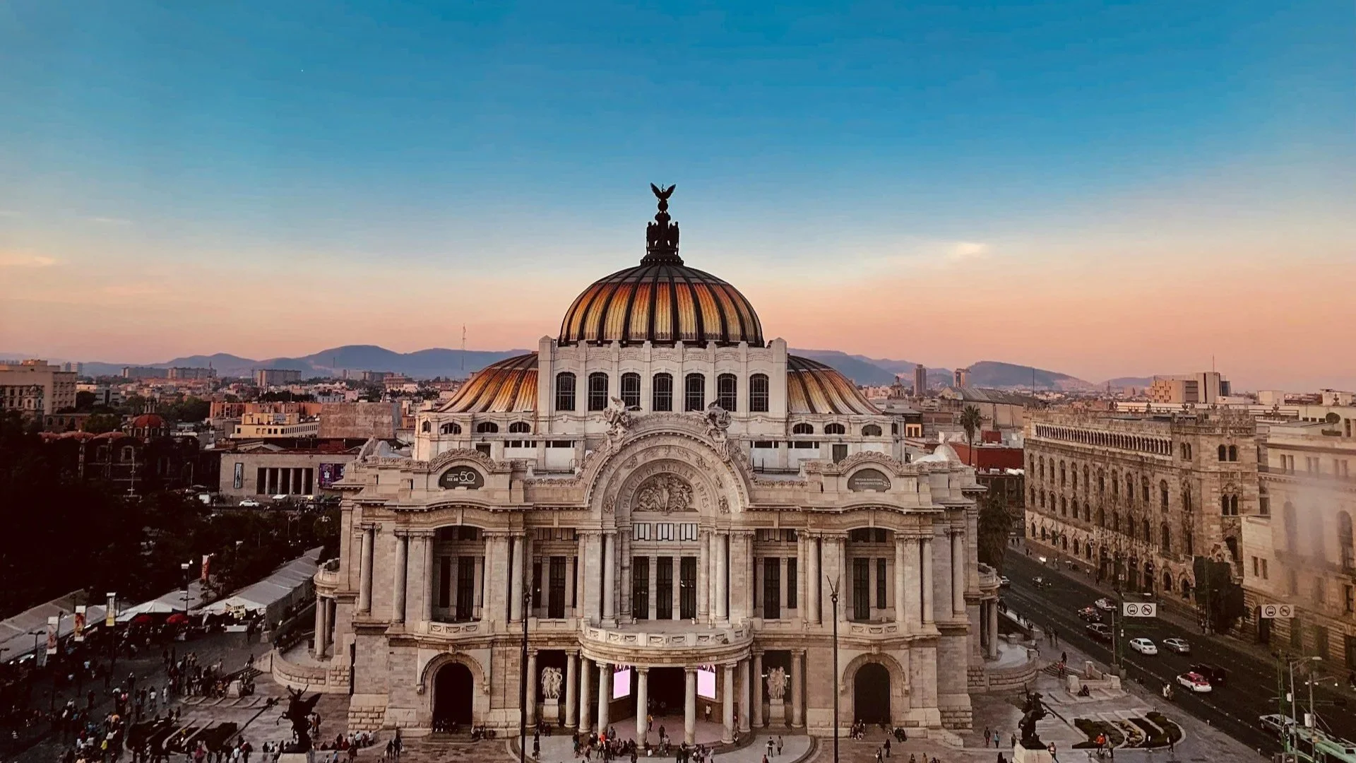 The Palacio de Bellas Artes in Mexico City at sunset, showing its white marble facade and golden-orange dome against a soft blue and pink sky.