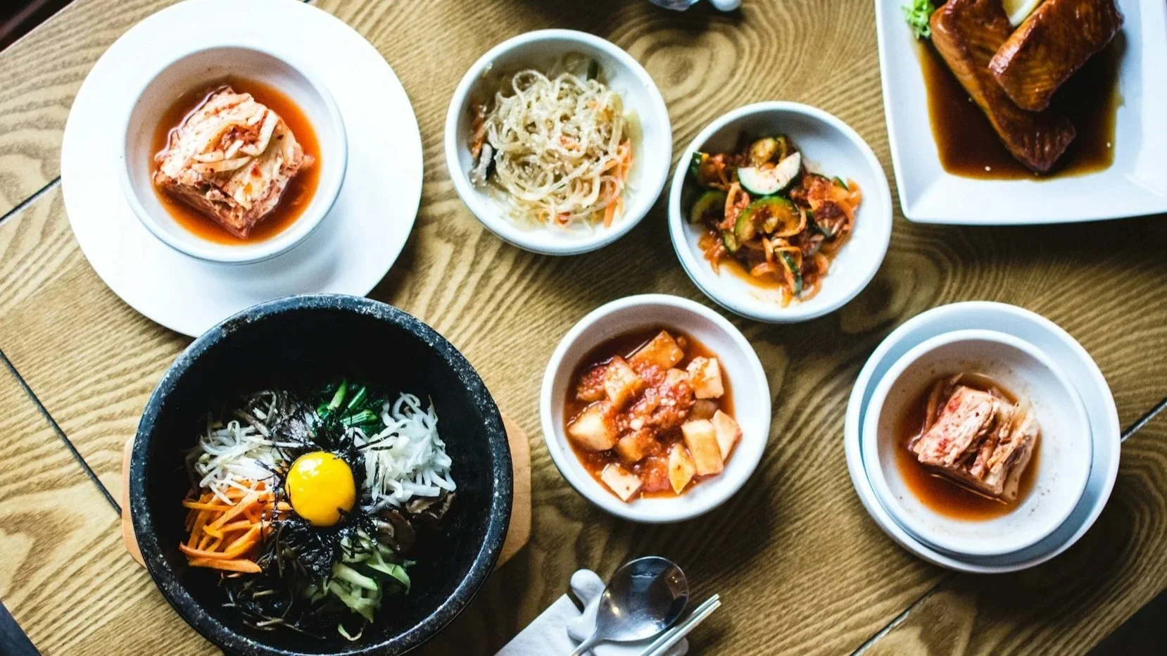 An overhead view of a traditional Korean meal on a wooden table, featuring a large stone bowl of bibimbap topped with a raw egg yolk, surrounded by various side dishes (banchan) like kimchi, radish, and sprouts.