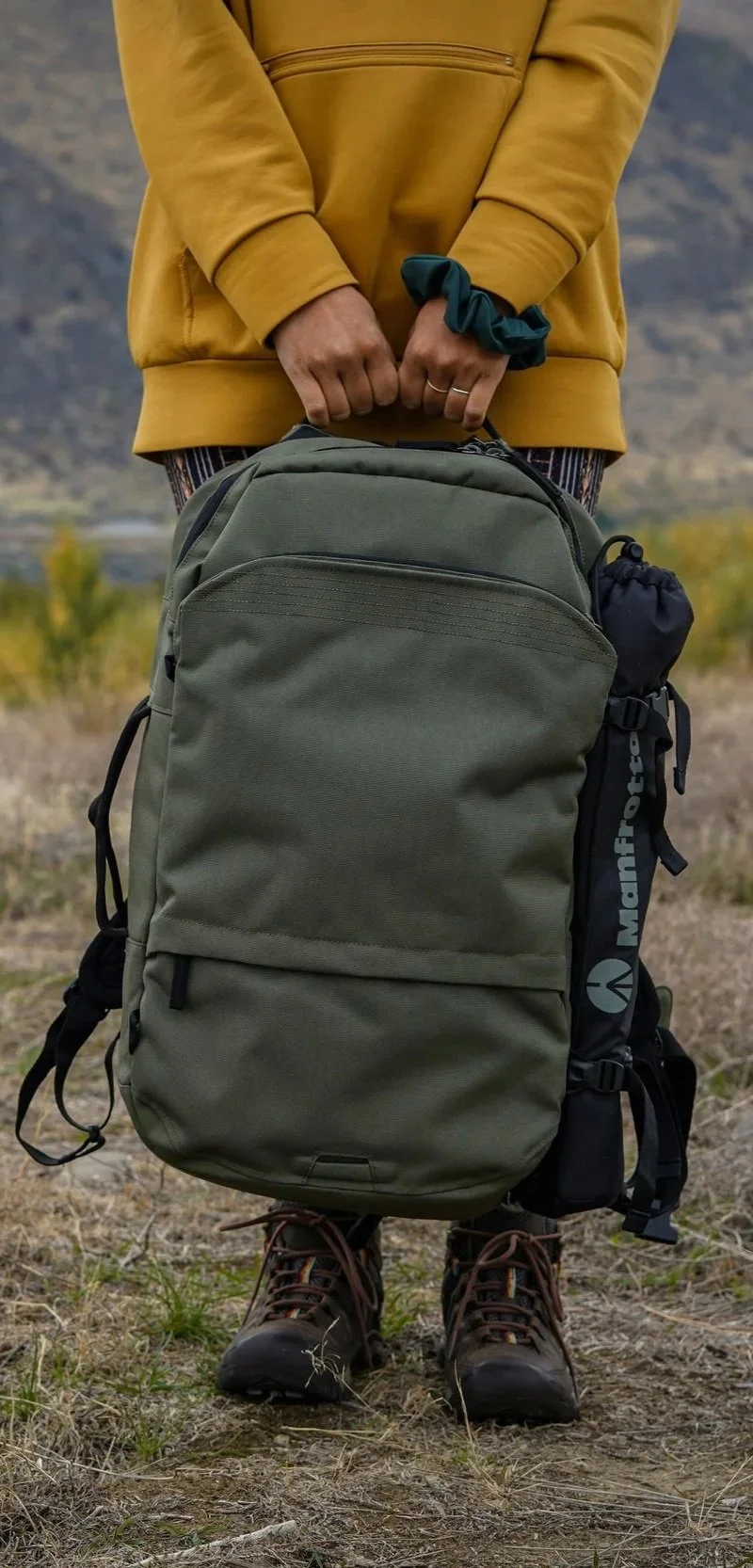 A cropped, waist-down shot of a person wearing a yellow hoodie and brown hiking boots, standing in a field with dry grass. The person is holding a large, olive-green backpack with two hands.