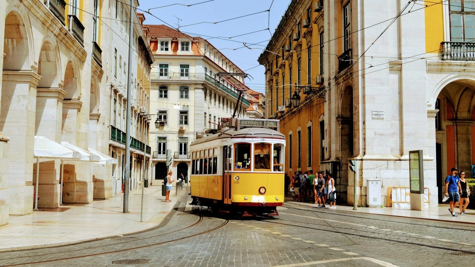 A classic yellow vintage tram traveling through a historic Lisbon street lined with grand, light-colored architecture.