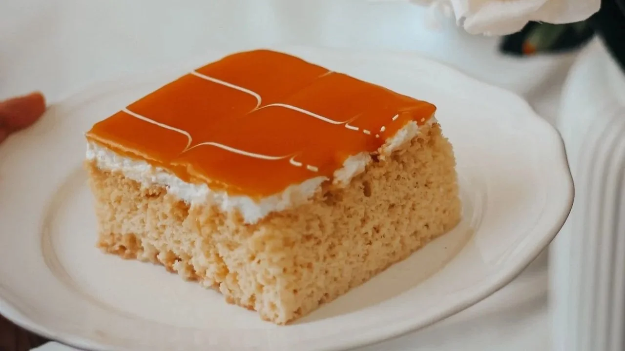 A slice of milk cake with caramel topping on a plate next to white roses and a lit candle.
