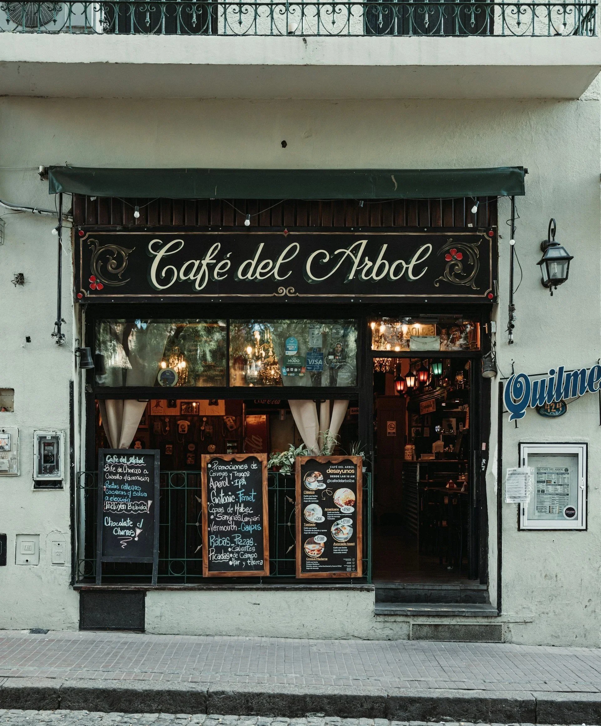 The storefront of a charming, vintage restaurant named 'Café del Arbol' features chalkboard menus detailing daily specials alongside an open doorway, all situated next to a cobblestone street.