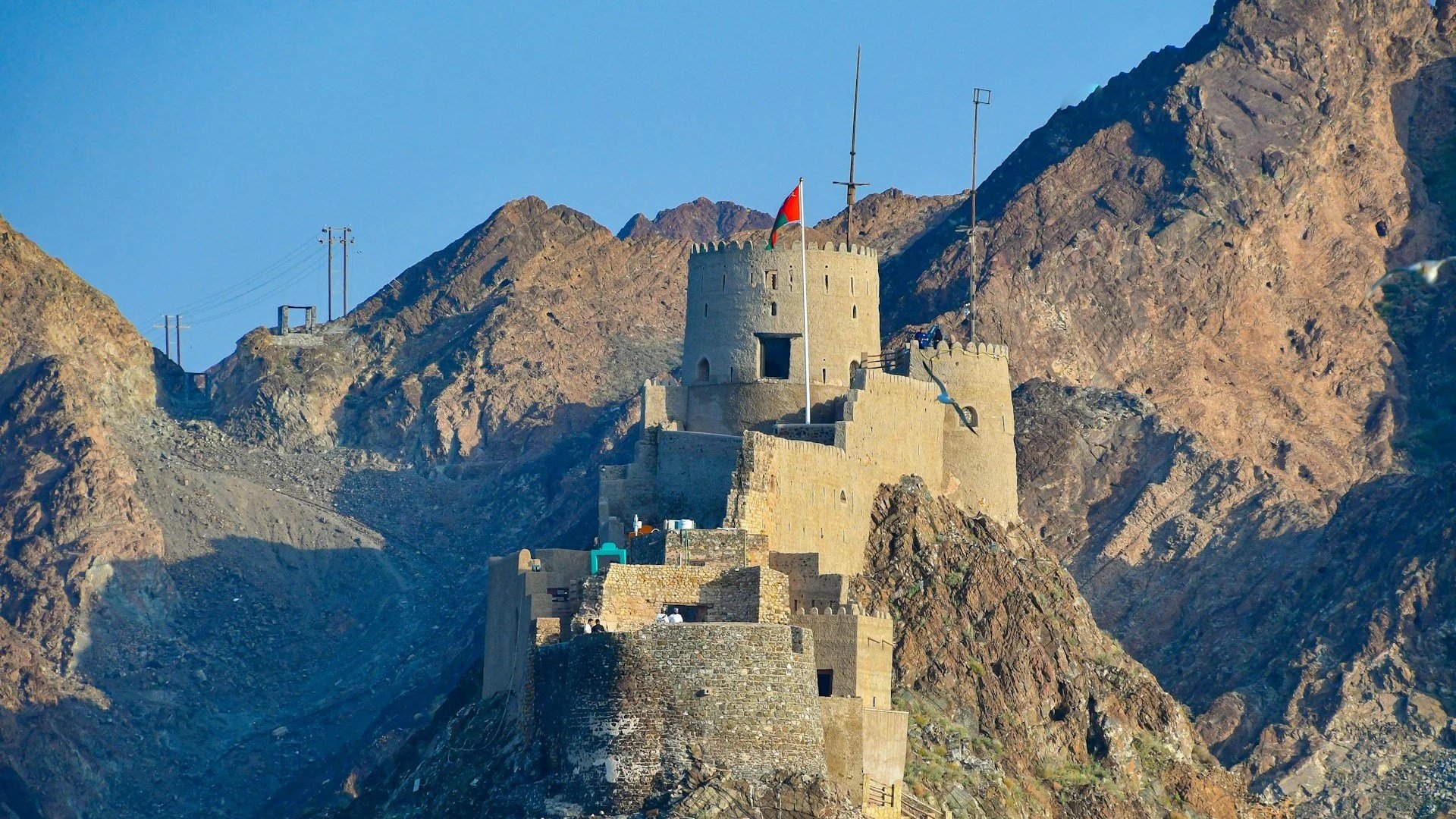 An ancient stone fortress with a central tower built into a rugged, steep mountain under a clear blue sky, with an Omani flag flying from its highest point.