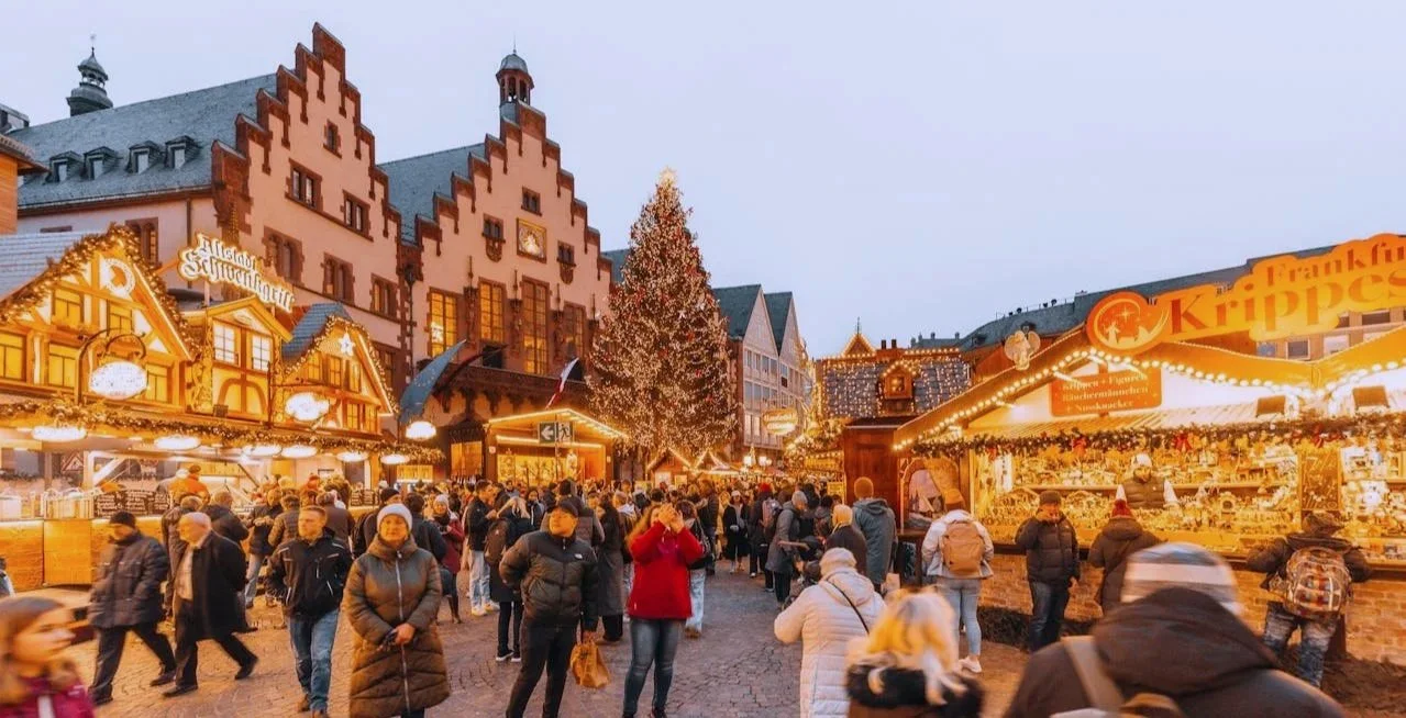 A vibrant, bustling Christmas market at night with glowing festive stalls, warm golden lights, and a crowd of people enjoying the holiday atmosphere in a historic town square.