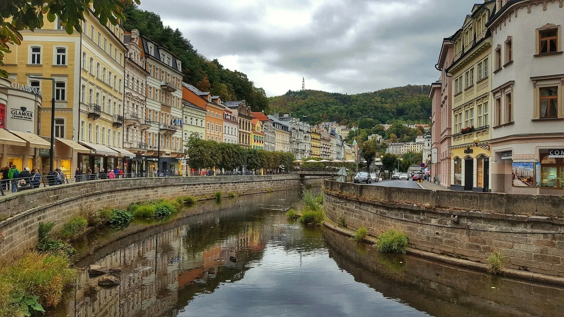 A scenic view of a canal lined with colorful, historic European-style buildings and lush green hills in the background.