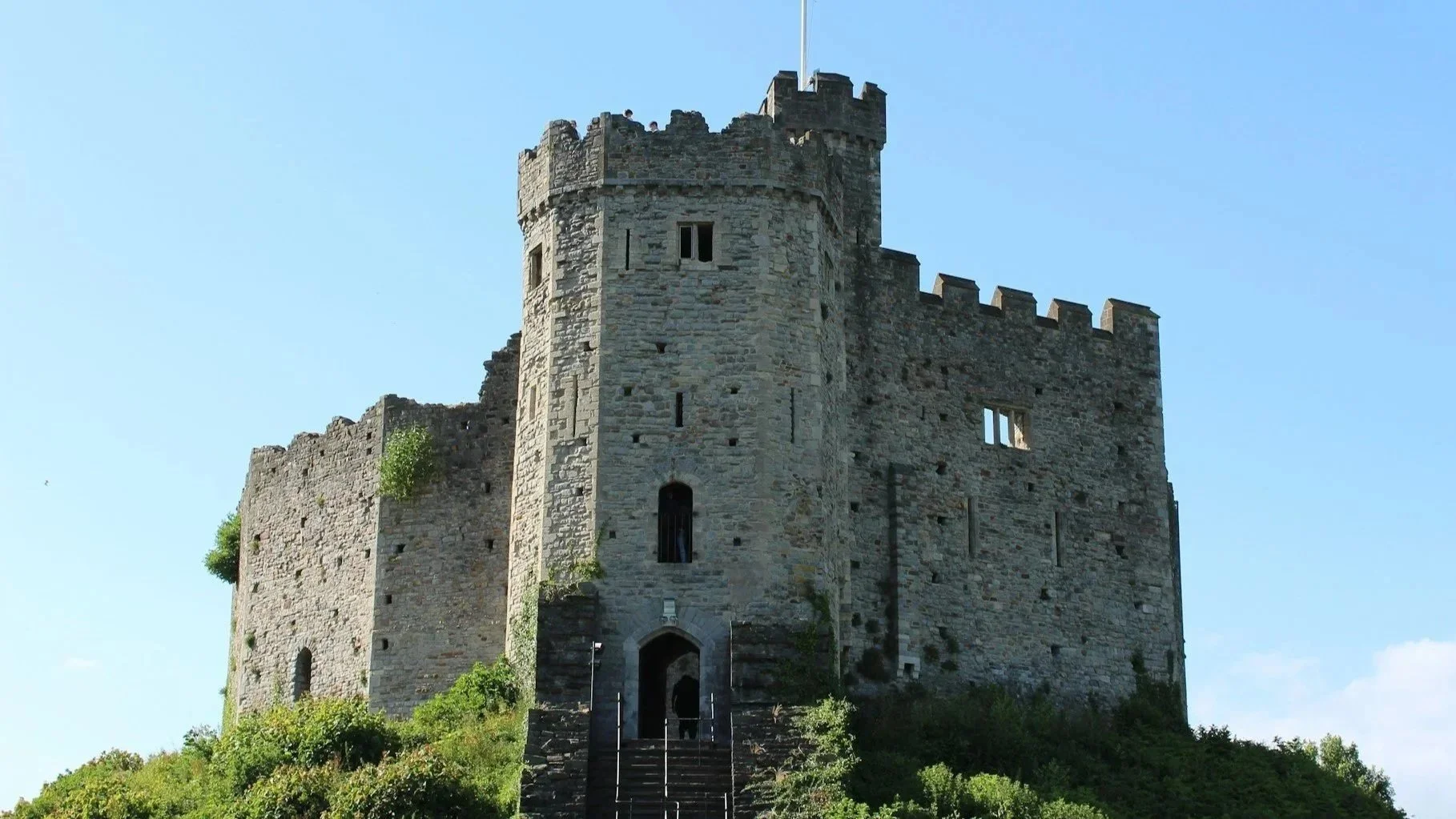 A historic stone castle tower sitting on a grassy mound with the Welsh flag flying from the top against a clear blue sky.