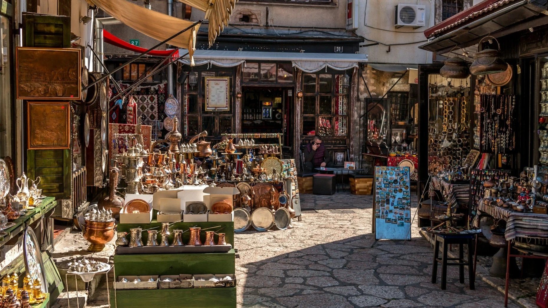 A cobblestone alley in an old-world market featuring shops filled with ornate copper crafts, coffee pots, and metal souvenirs under a tan awning.