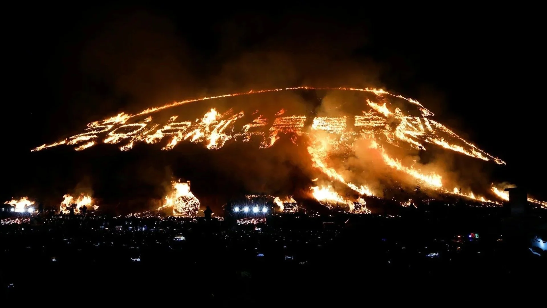 Large-scale fire burning in patterns and Korean characters across a dark hillside at night.