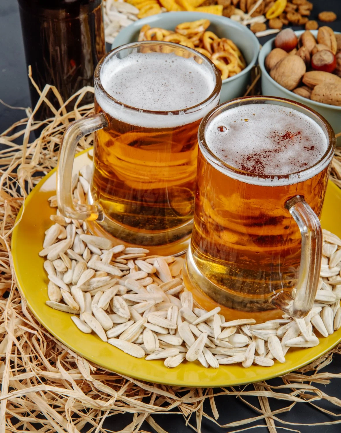 Two foaming mugs of beer on a yellow plate surrounded by sunflower seeds, pretzels, and mixed nuts.
