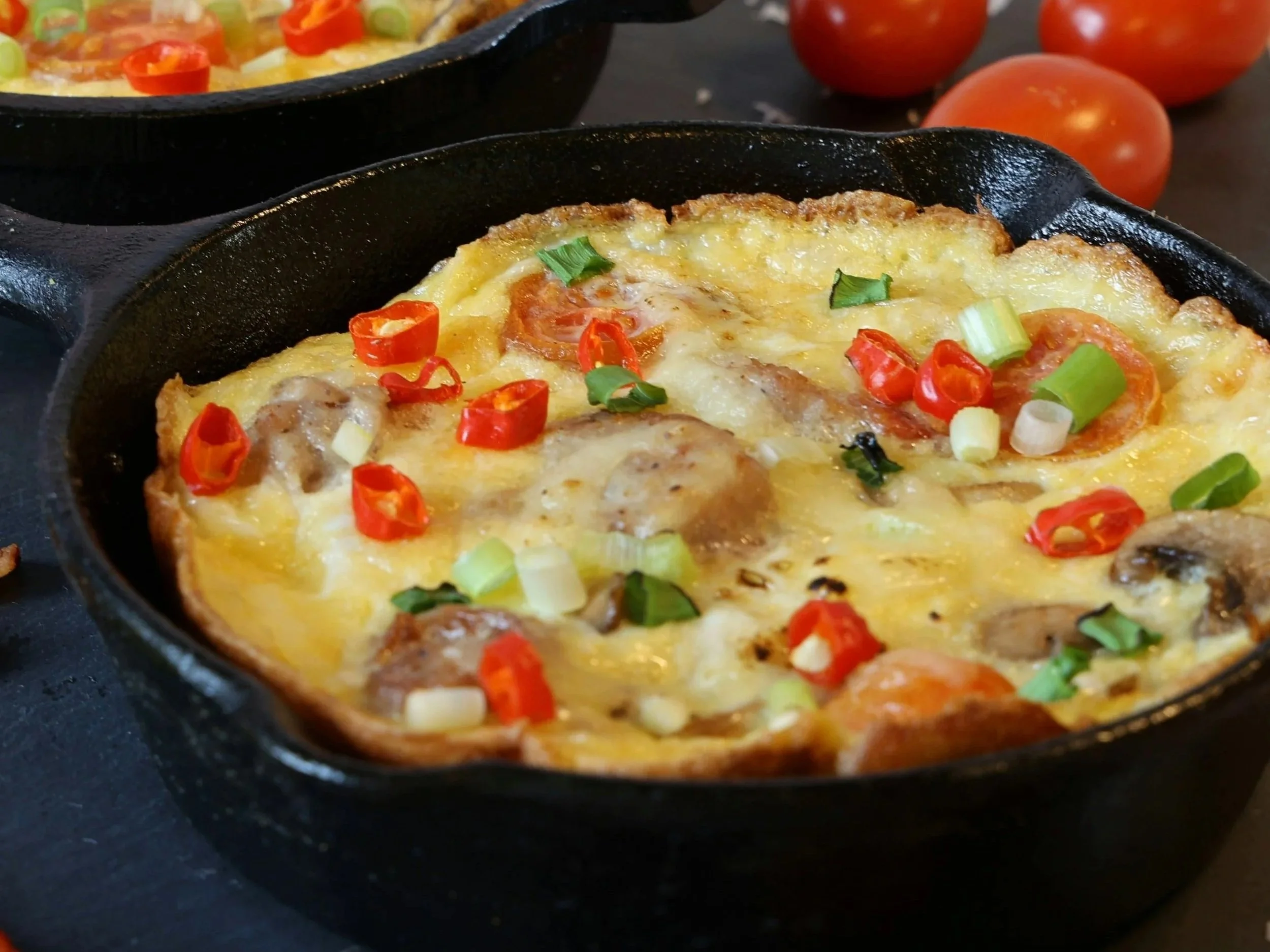 Two small cast-iron skillets filled with golden-brown frittatas topped with sliced red chilies, green onions, and mushrooms, sitting on a dark surface next to fresh tomatoes.