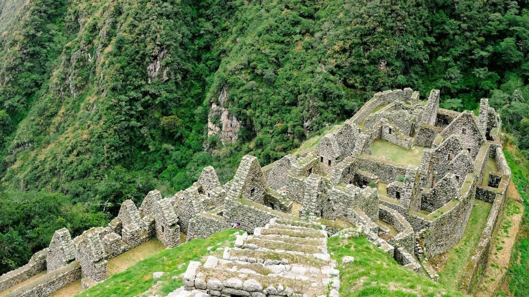 An overhead view of ancient stone structures and agricultural terraces nestled against a lush, green mountainside.