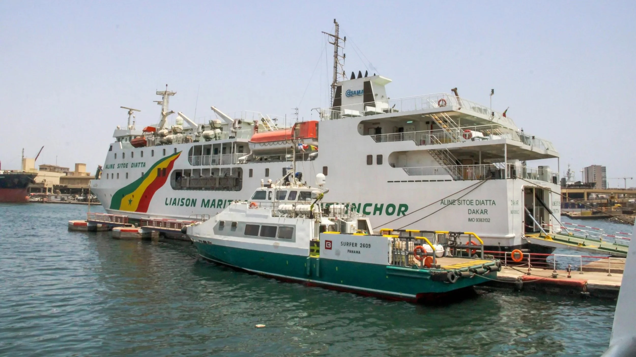 A large white passenger ferry named "Aline Sitoe Diatta" from Dakar docked at a port next to a smaller green and white service boat.