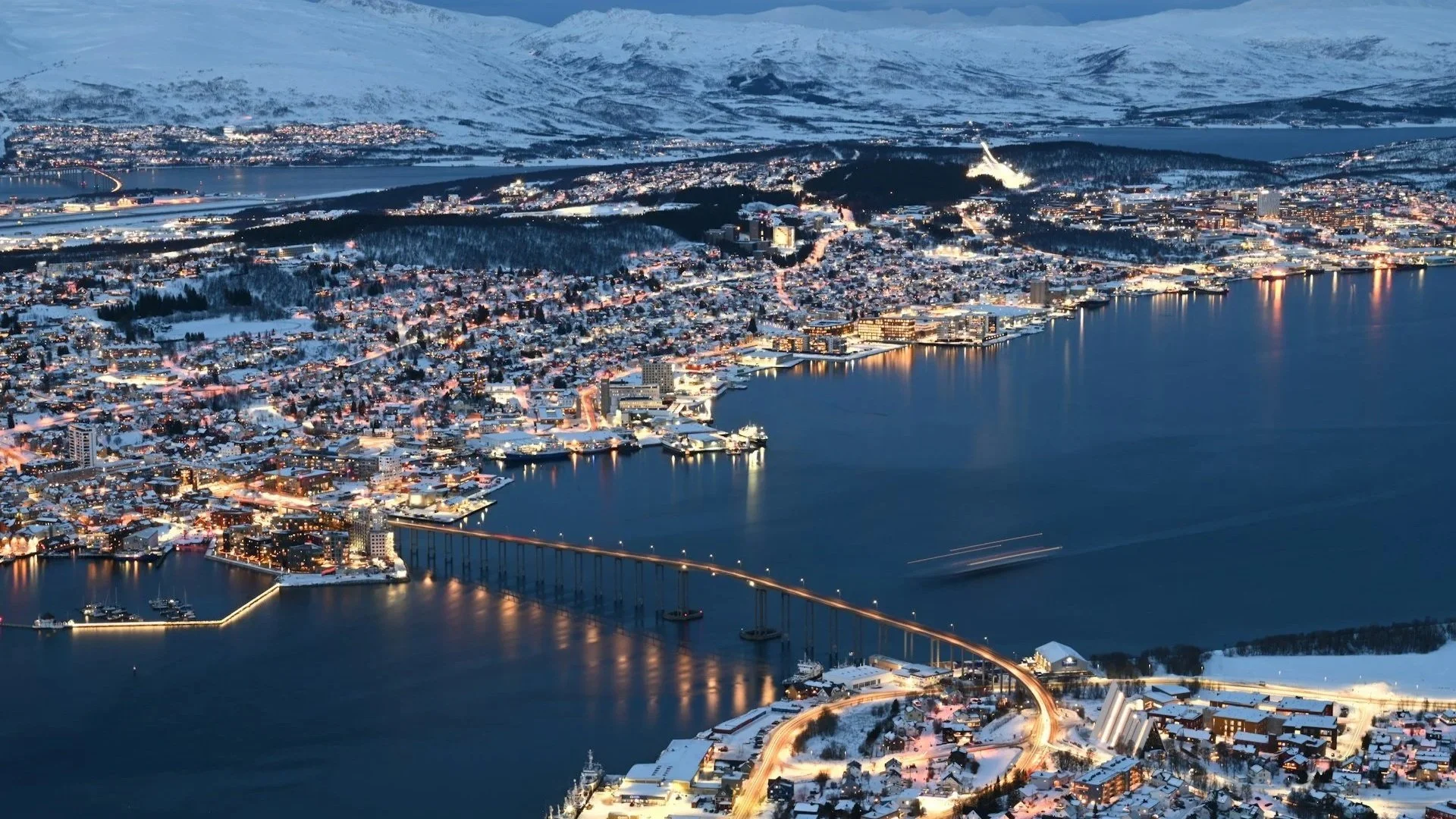 An aerial night view of a snow-covered coastal city in Norway, featuring a long bridge and glowing city lights.