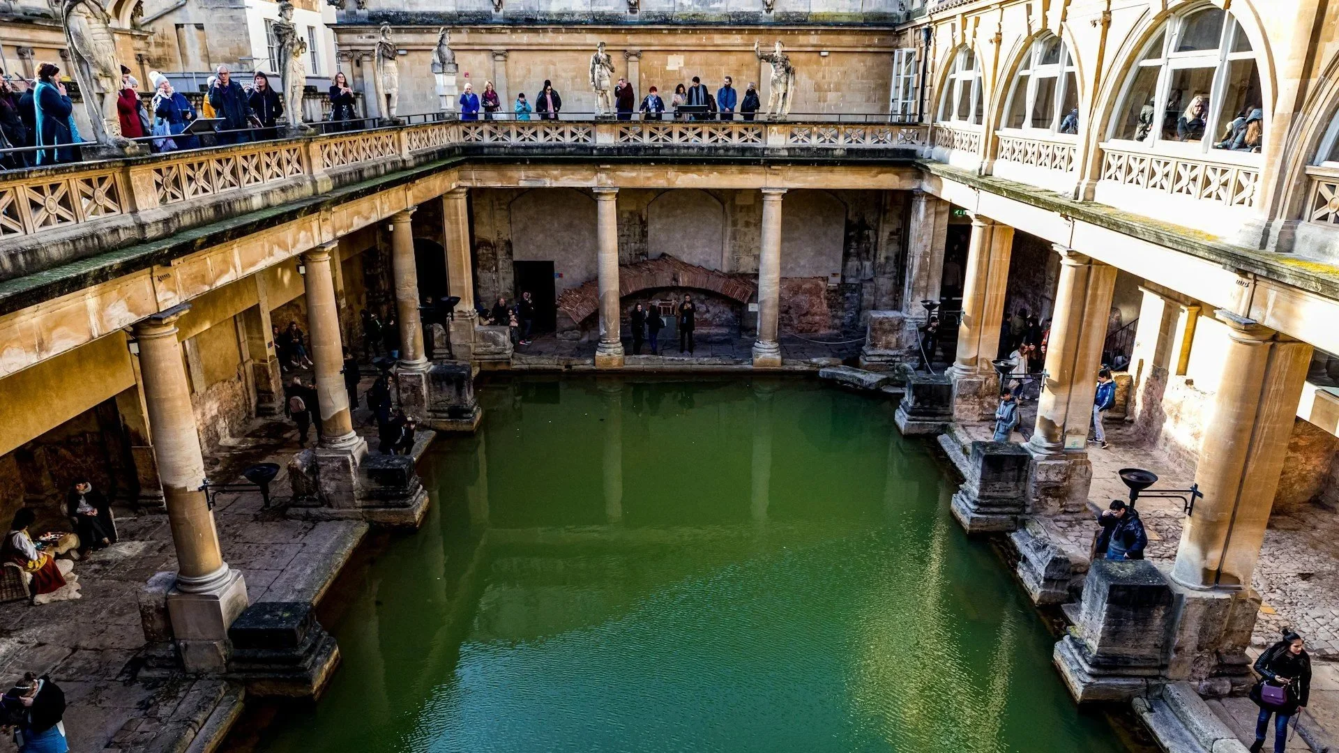 An overhead view of the Great Bath at the Roman Baths in Bath, England, showing the green geothermal water and surrounding ancient stone columns.