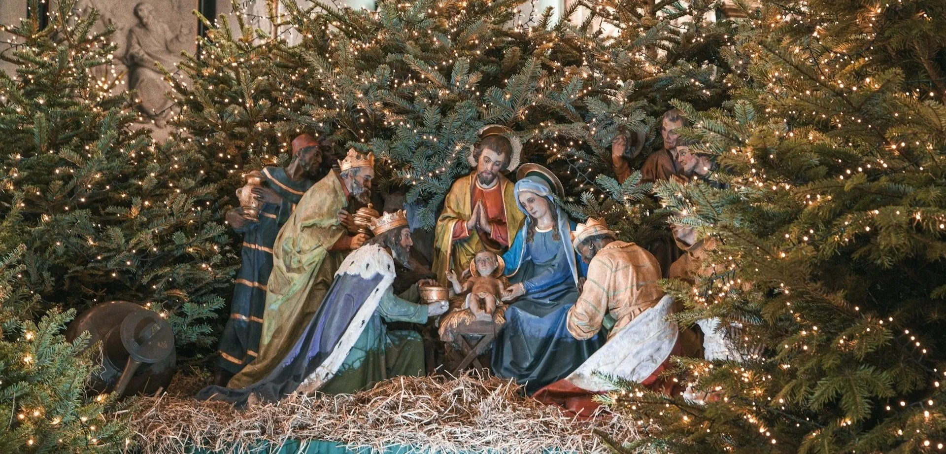 A traditional nativity scene display surrounded by small evergreen trees with twinkling lights.